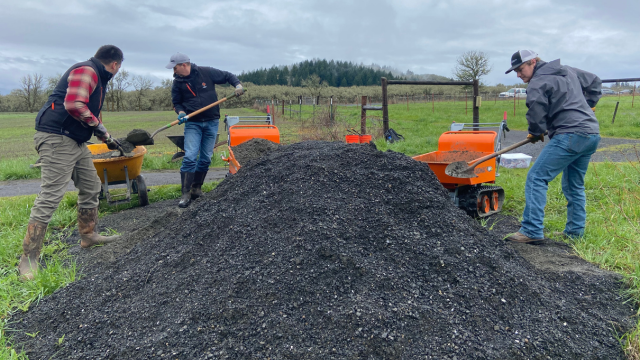 Three men shovel gravel from a large pile into bright colored wheelbarrows