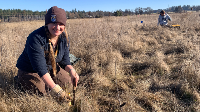 A woman in a warm hat digs in the ground in an open prairie